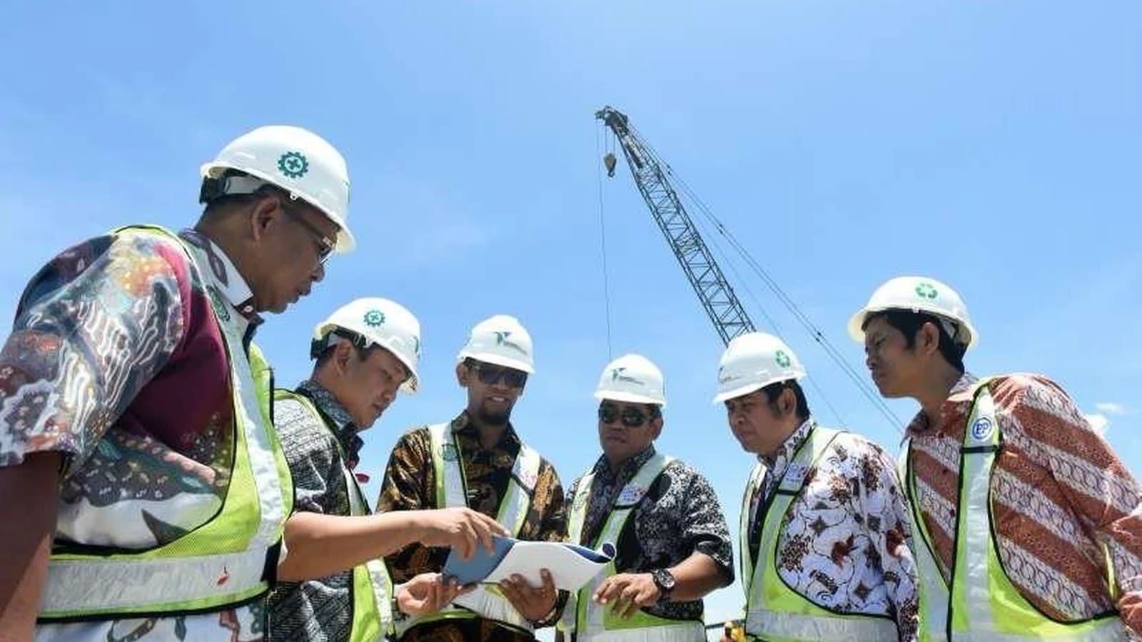 Enam orang mengenakan helm proyek putih dan rompi reflektif di atas kemeja batik meninjau sebuah dokumen bersama di luar ruangan, dengan crane di bawah langit biru di belakang mereka.